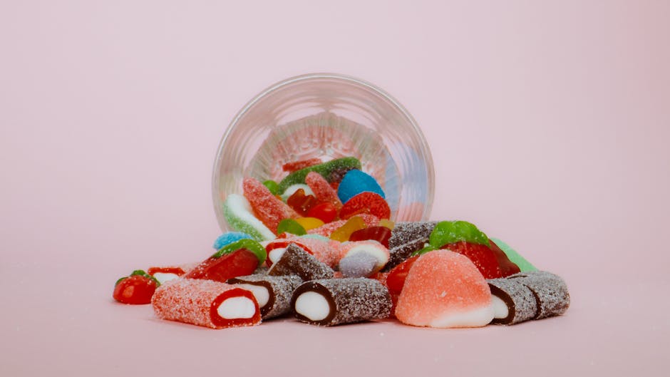 An assortment of colorful gummy candies spilling from a clear glass on a pink background.