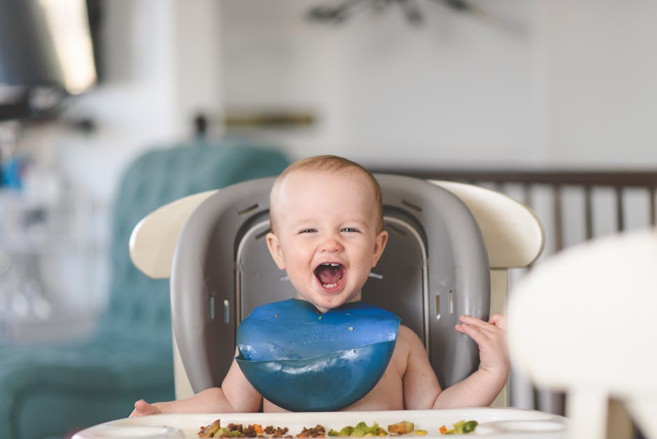 Happy baby laughing in a high chair during mealtime, wearing a blue bib.
